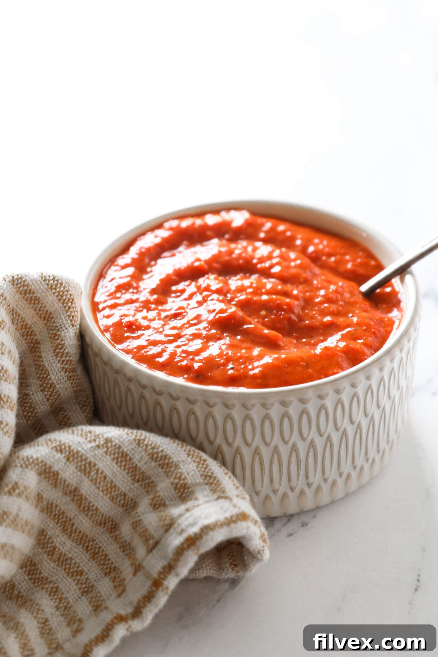 A spoonful of creamy red pepper sauce being lifted from a white bowl, showcasing its smooth, thick texture and vibrant red color.