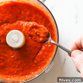 Overhead image of a spoonful of red pepper sauce over a food processor, highlighting the sauce's texture.