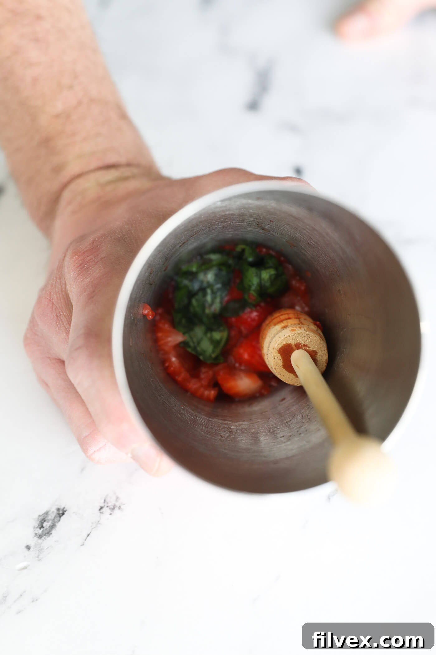 A muddler actively crushing fresh strawberries and basil leaves within a cocktail shaker, demonstrating the release of juices and aromatic oils from the ingredients.