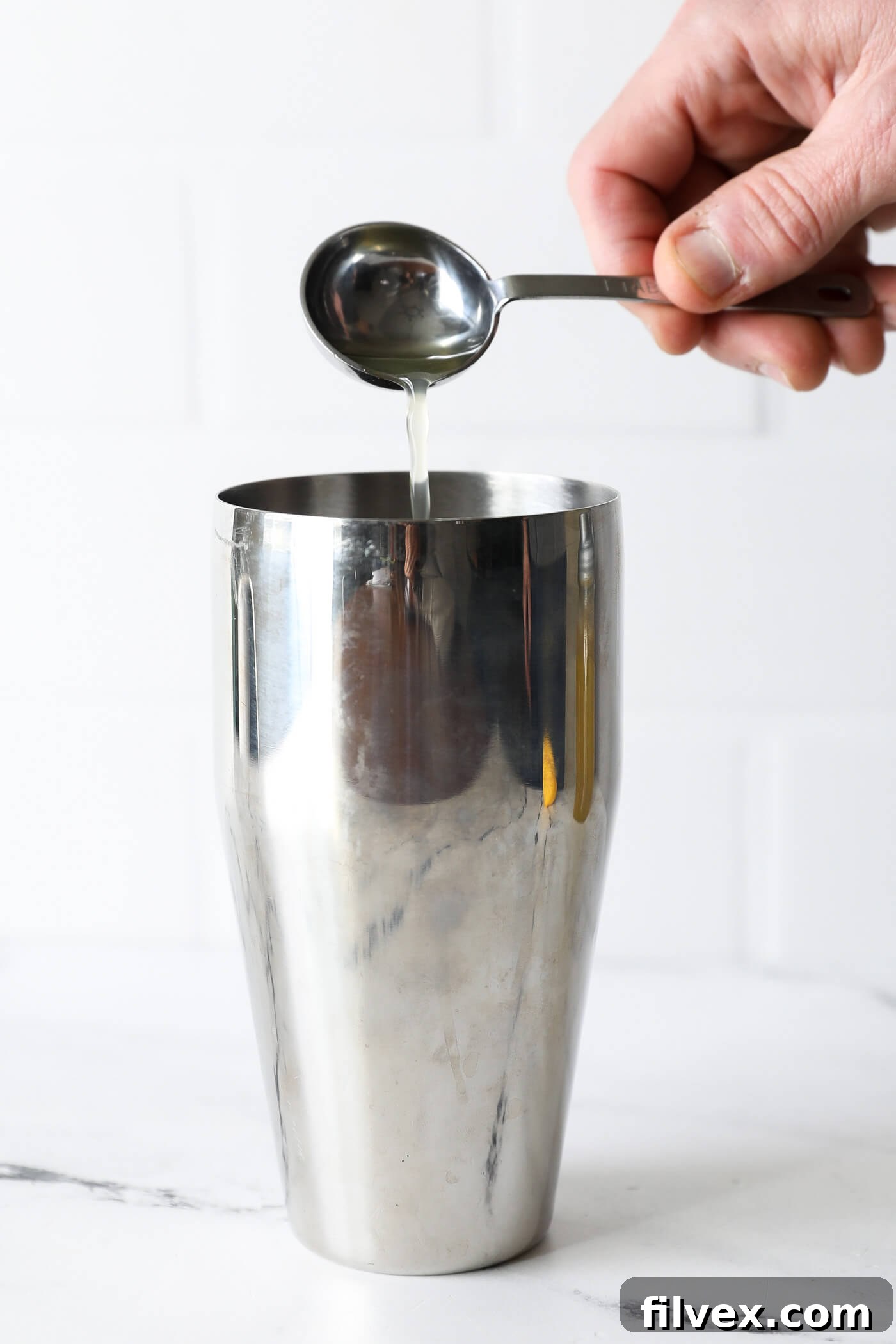 A stream of golden honey simple syrup is being poured into the cocktail shaker, joining the muddled strawberries and basil.