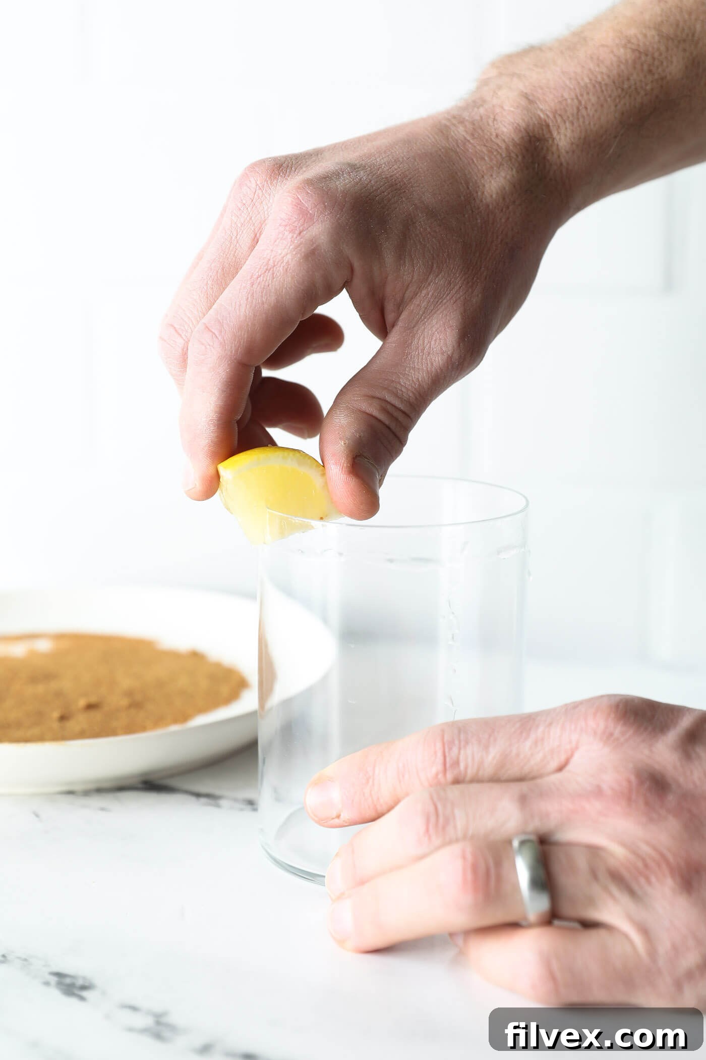 A hand holds a lemon wedge, moistening the rim of a clear glass in preparation for an optional sugar coating, creating a sticky surface.