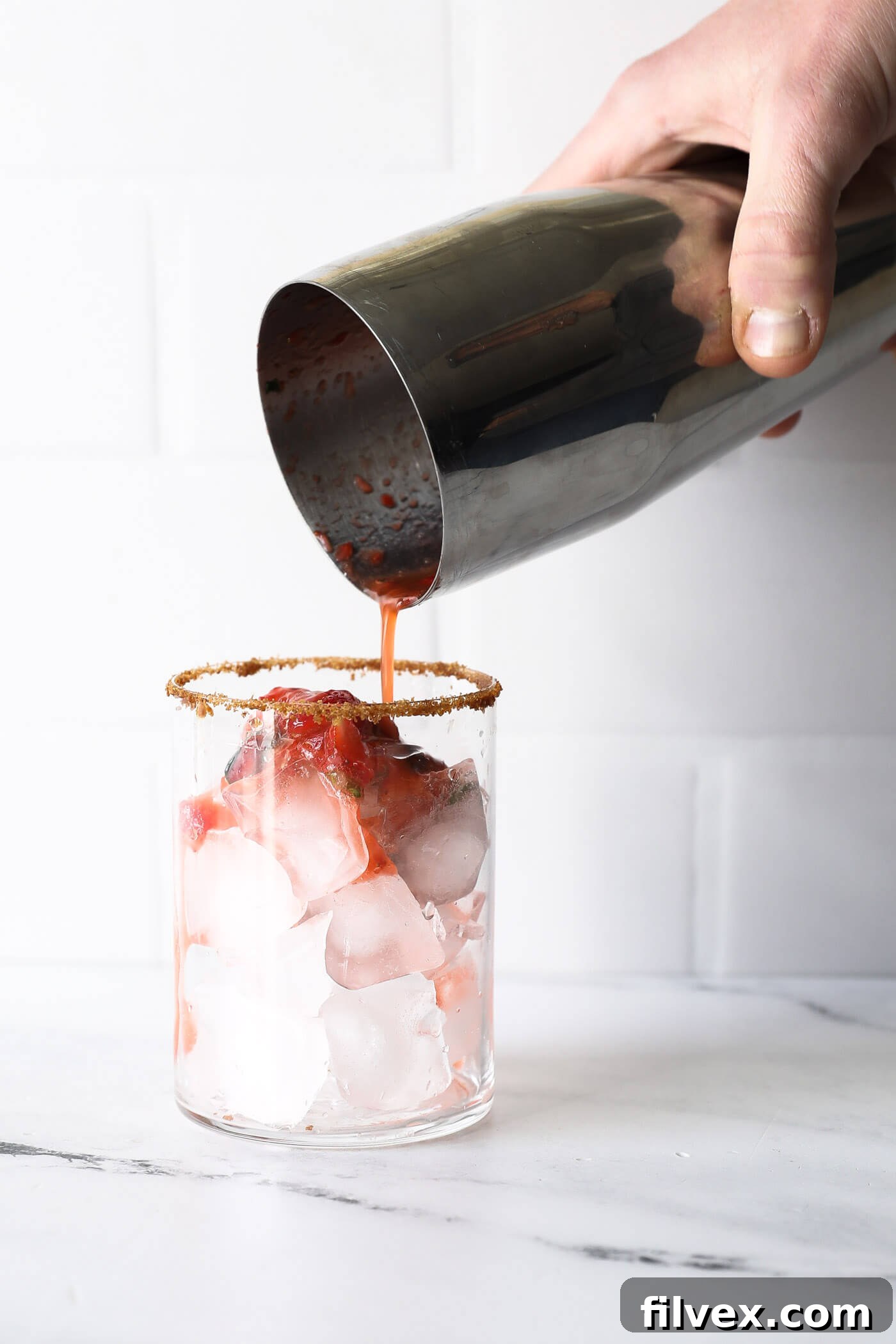 A prepared glass with a sugar-coated rim is being filled with clear ice cubes, creating the foundation for the chilled mocktail mixture.