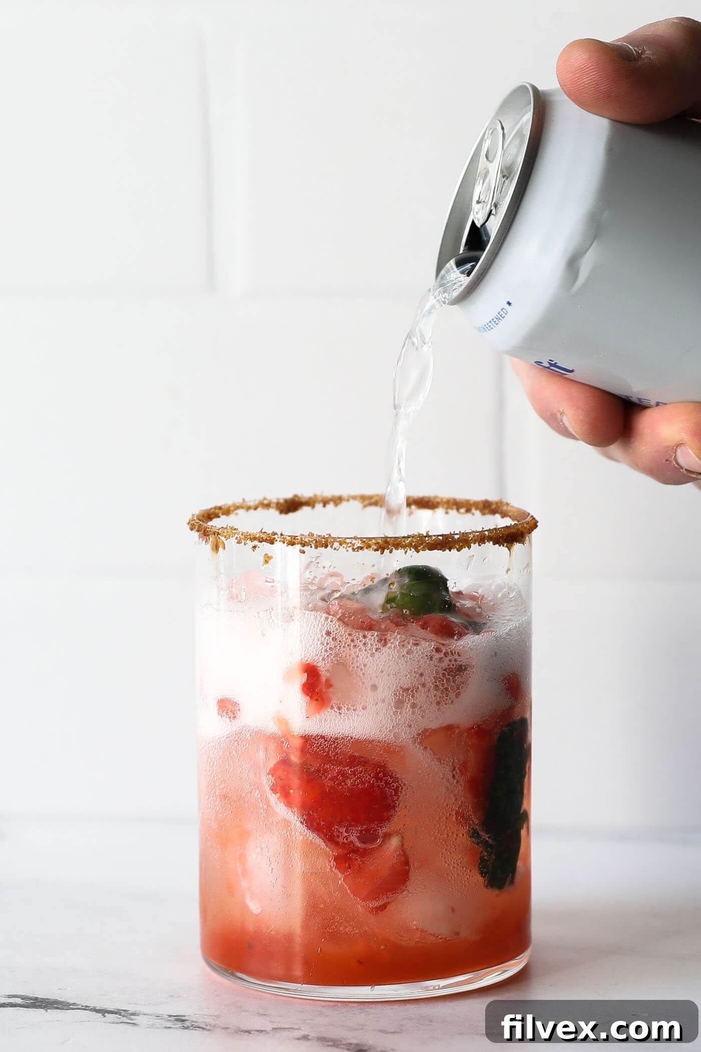 A hand pours sparkling water into a glass already containing the vibrant strawberry mixture and ice, creating the refreshing fizz of the mocktail.