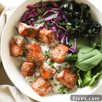 Cooked salmon bites served over rice, greens, and purple cabbage in a bowl, garnished with sesame seeds