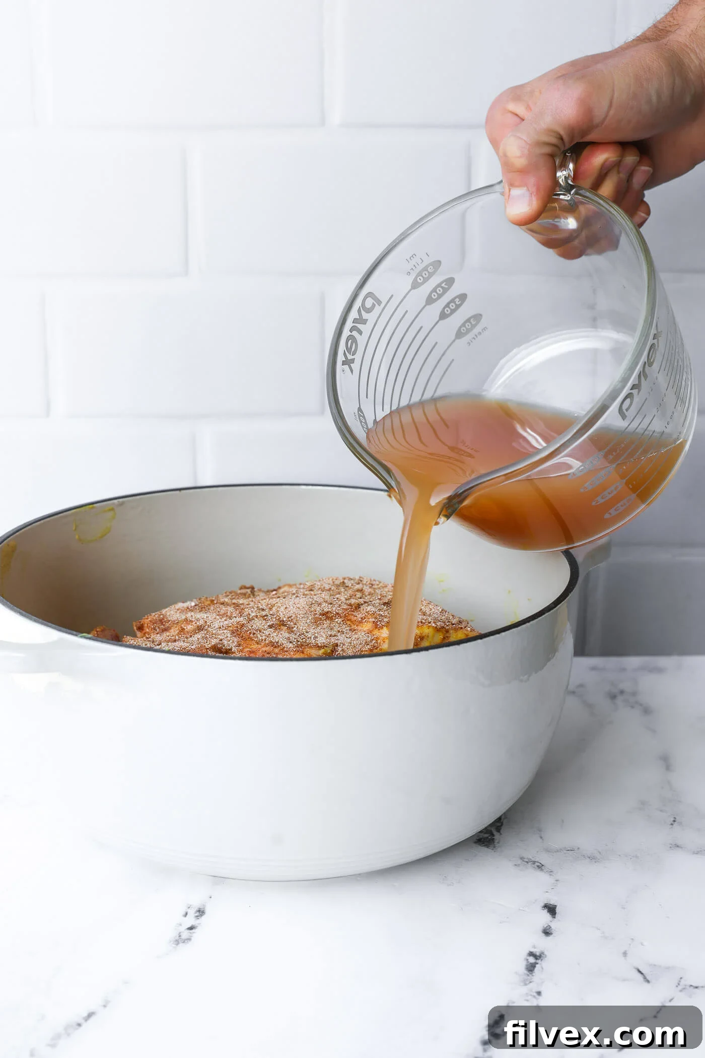 Apple juice being poured into a Dutch oven around the seasoned pork shoulder.