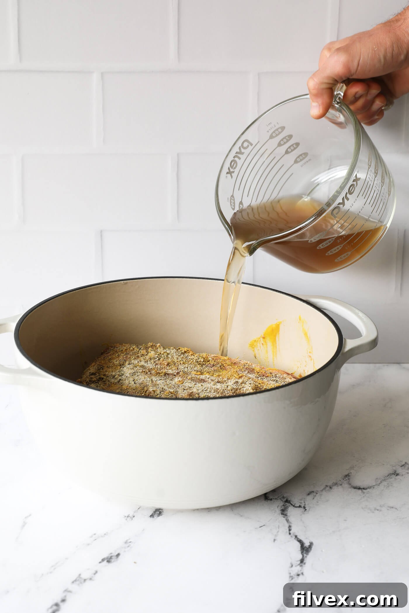 Pouring beef broth into a Dutch oven around the seasoned pot roast