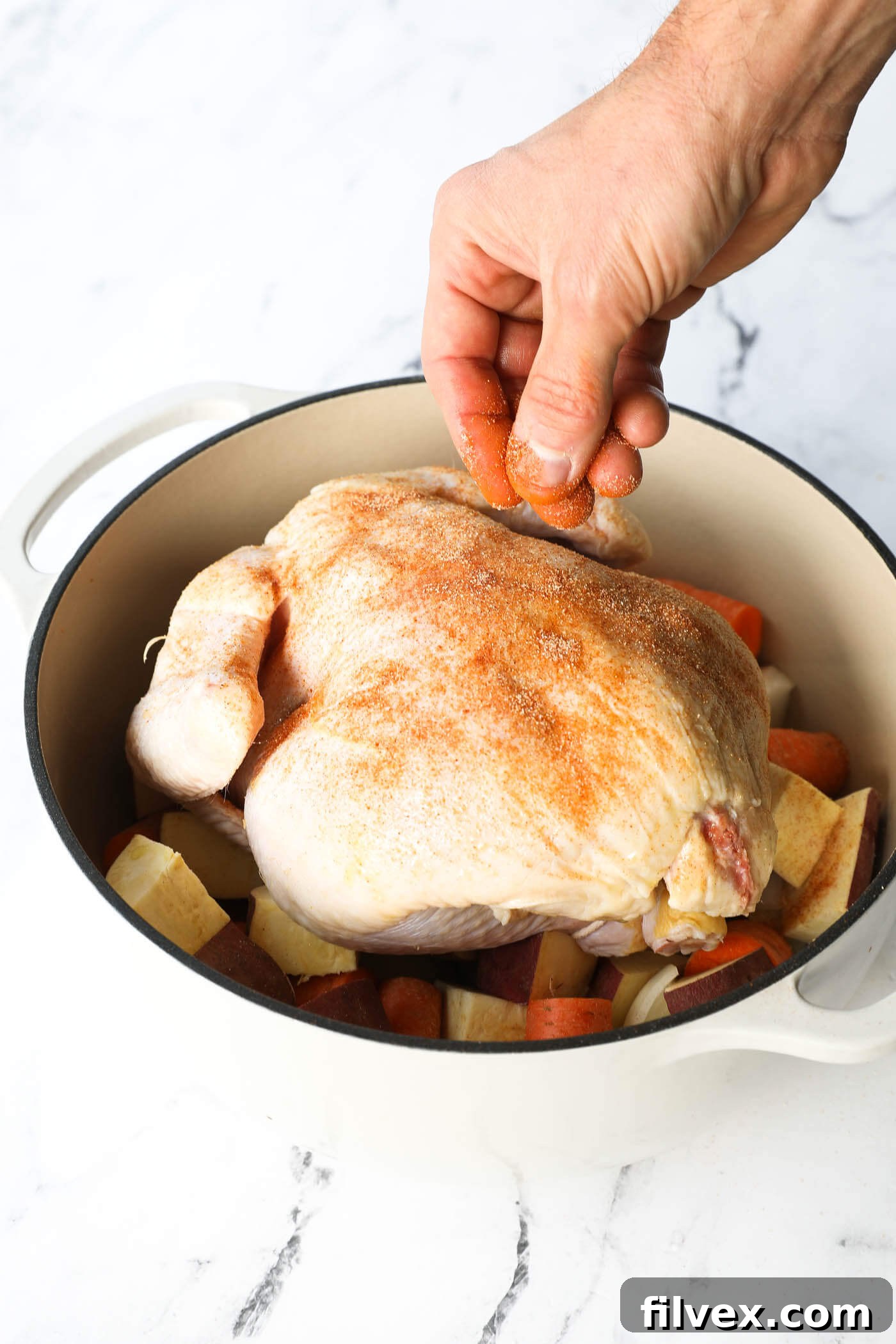 Angled image of a hand sprinkling seasonings on a raw chicken in a dutch oven before cooking.