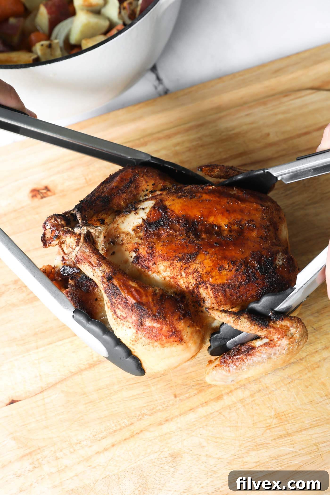 Someone using two tongs to remove the cooked whole chicken from the dutch oven and placing on a cutting board to rest.
