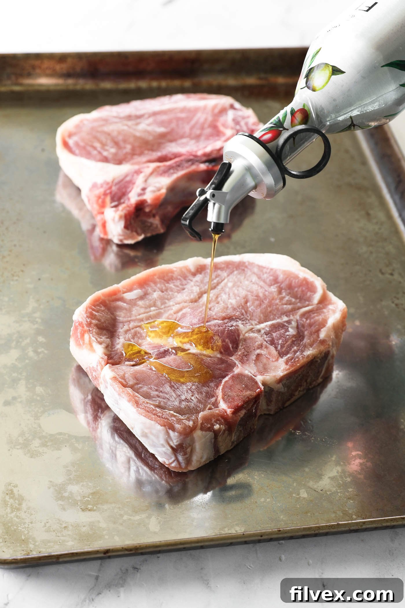 A stream of olive oil being drizzled over raw, thick-cut pork chops arranged on a metal baking sheet.