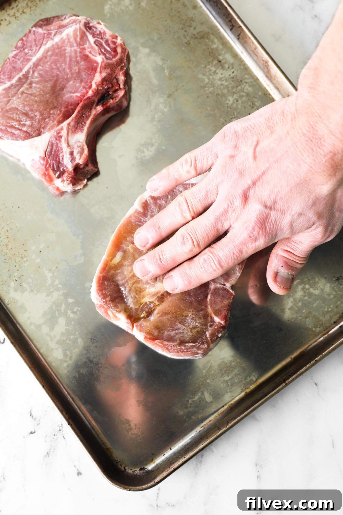 A hand spreading olive oil evenly over the surface of a pork chop using a brush, ensuring full coverage.