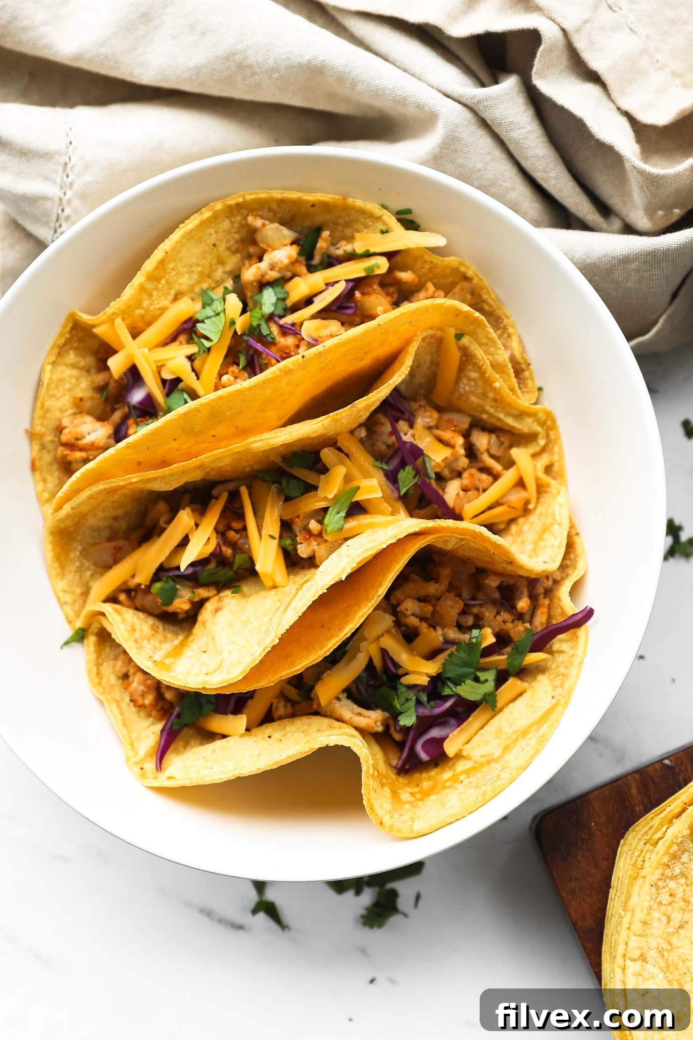 Overhead close up image of three tacos in a bowl. Ground chicken mixture, chopped cabbage, cilantro and shredded cheese on top.