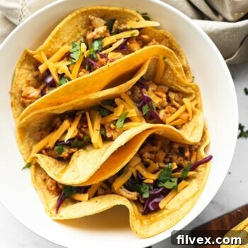 Overhead close up image of three tacos in a bowl. Ground chicken mixture, chopped cabbage, cilantro and shredded cheese on top.