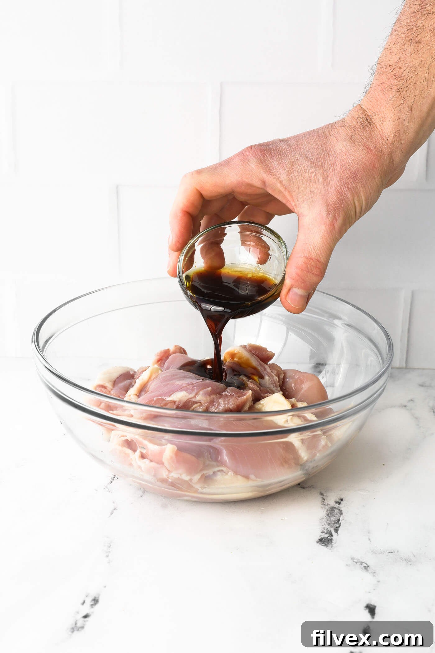 Pouring sauce over raw chicken thighs in a glass bowl. Uncooked chicken thighs being gently coated in a savory liquid mixture within a large glass mixing bowl, preparing them for seasoning.