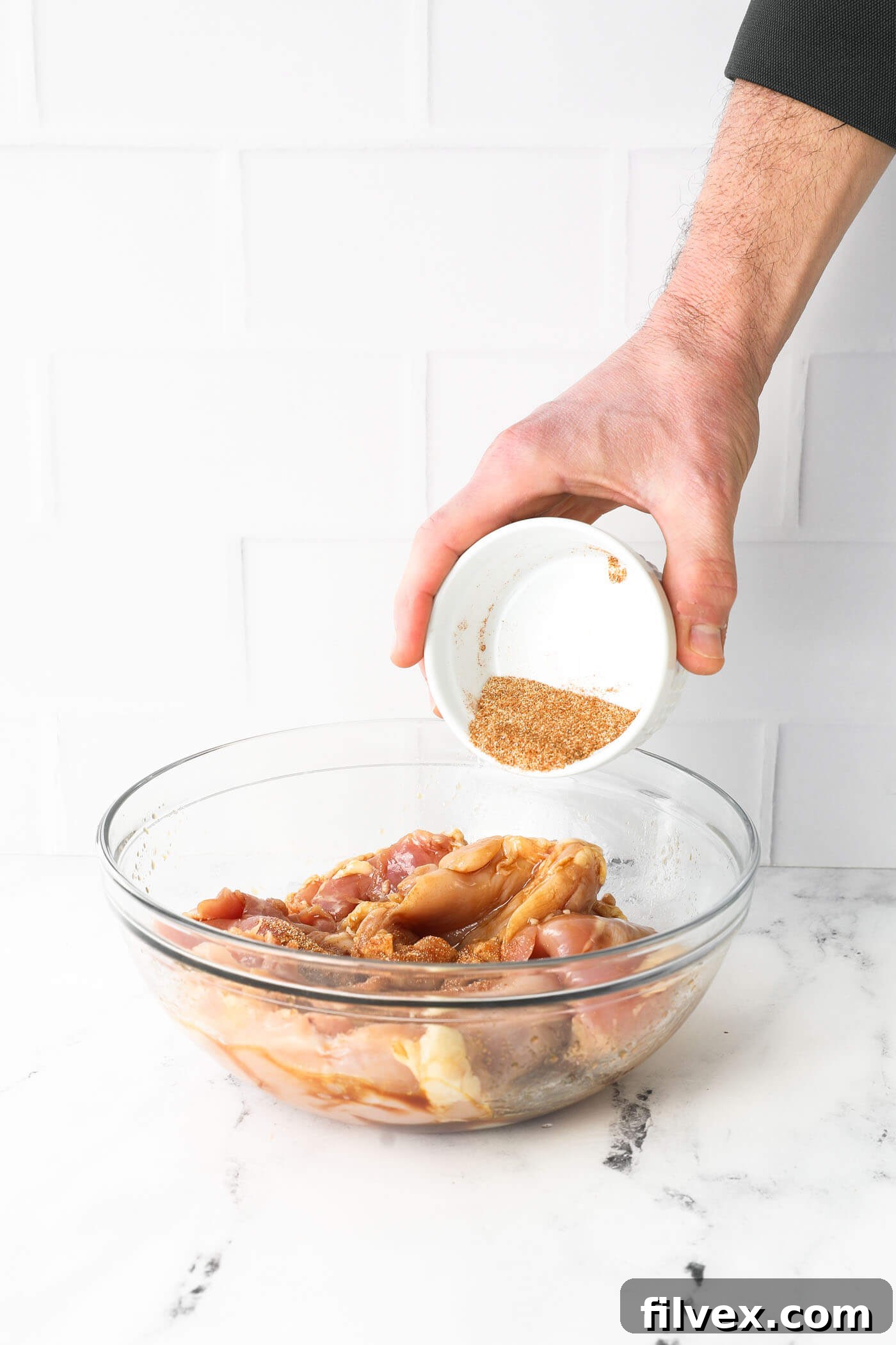 Pouring seasoning mix over the coated chicken in a glass bowl. A generous amount of the aromatic spice mixture being sprinkled evenly over the sauced chicken thighs in a glass bowl.