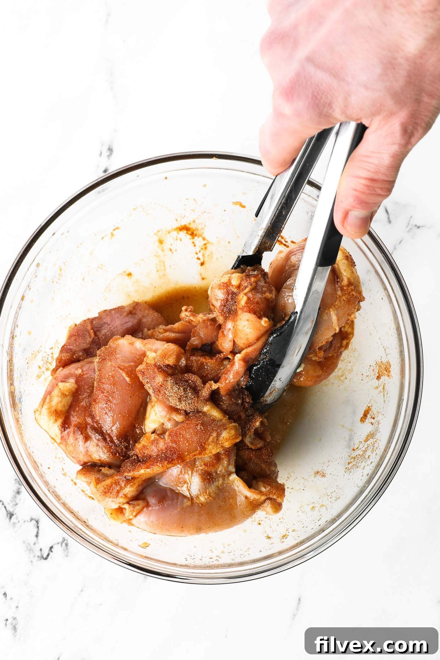 Using tongs to mix the seasonings onto the sauce coated chicken thighs in a glass bowl. Tongs mixing the chicken thighs to ensure the dry seasoning is thoroughly combined with the liquid coating.