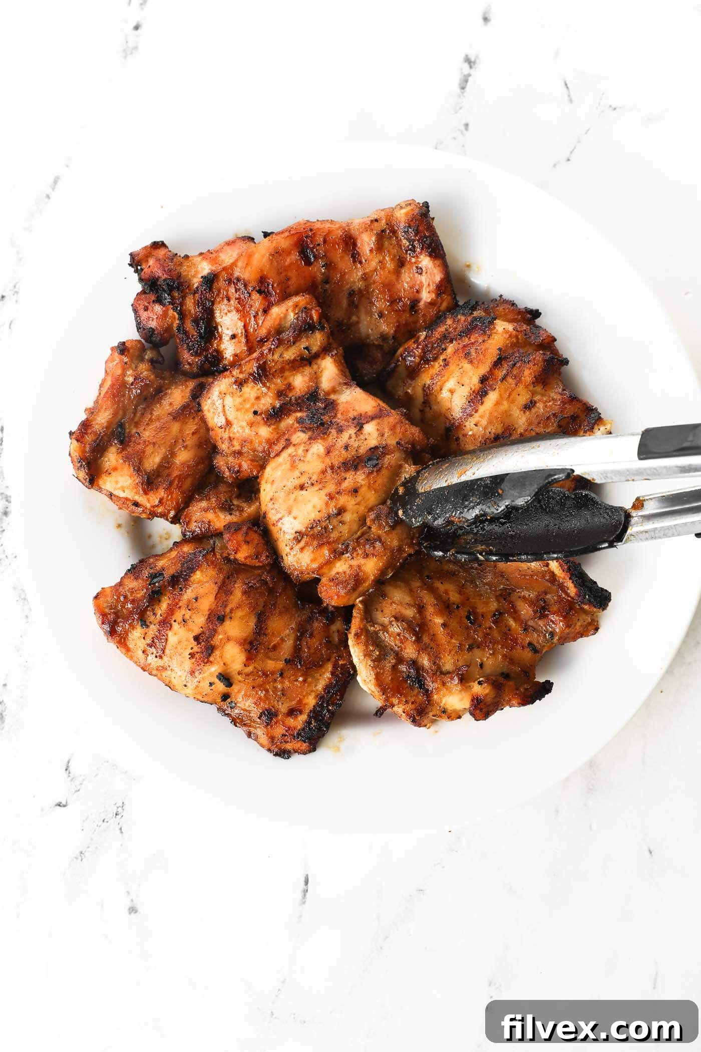 Adding cooked chicken thighs to a plate with tongs. Freshly grilled, golden-brown chicken thighs being transferred from the hot grill to a clean serving plate using tongs, ready for a brief rest before enjoying.