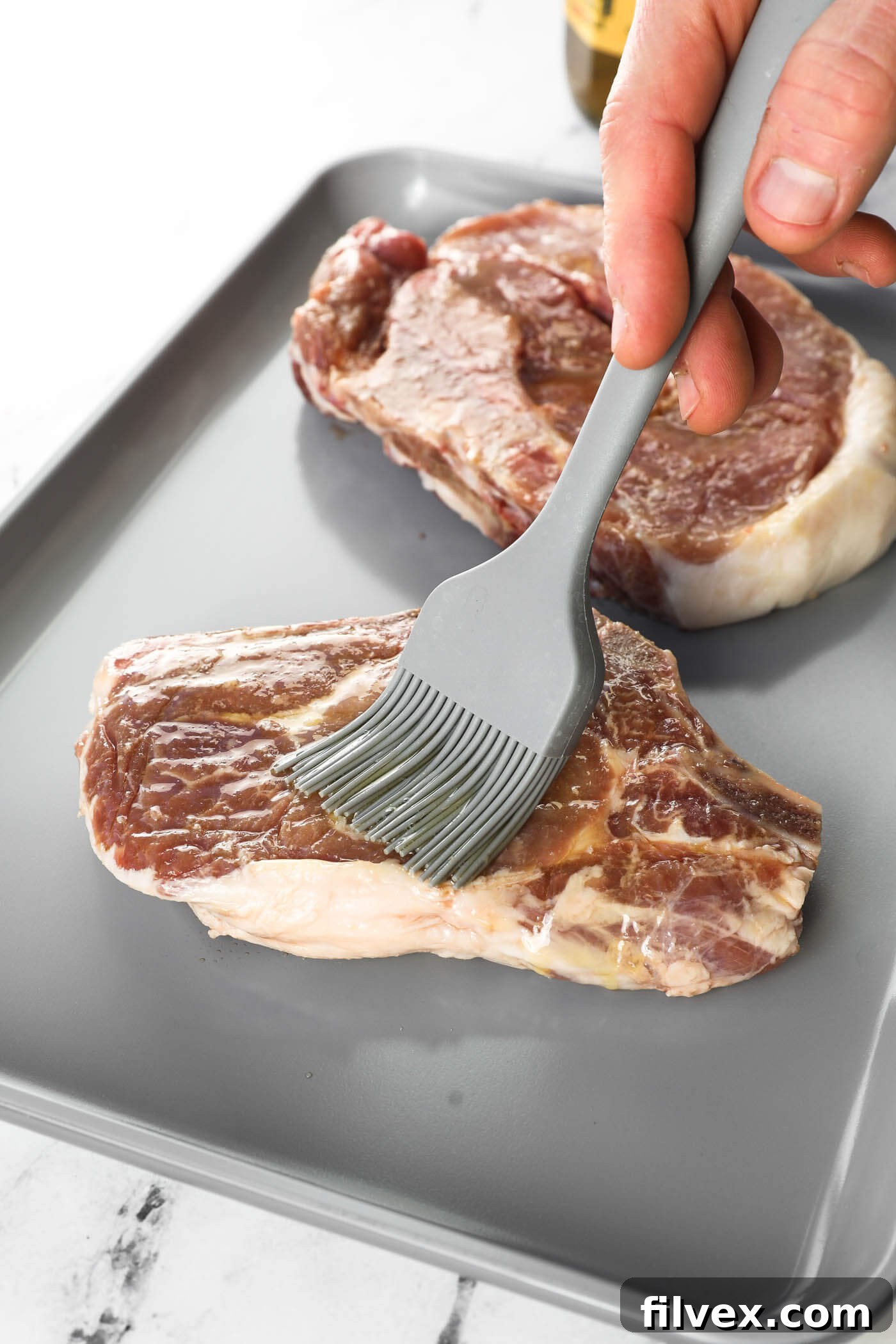 A raw bone-in pork chop being brushed with olive oil on a cutting board, preparing it for seasoning.