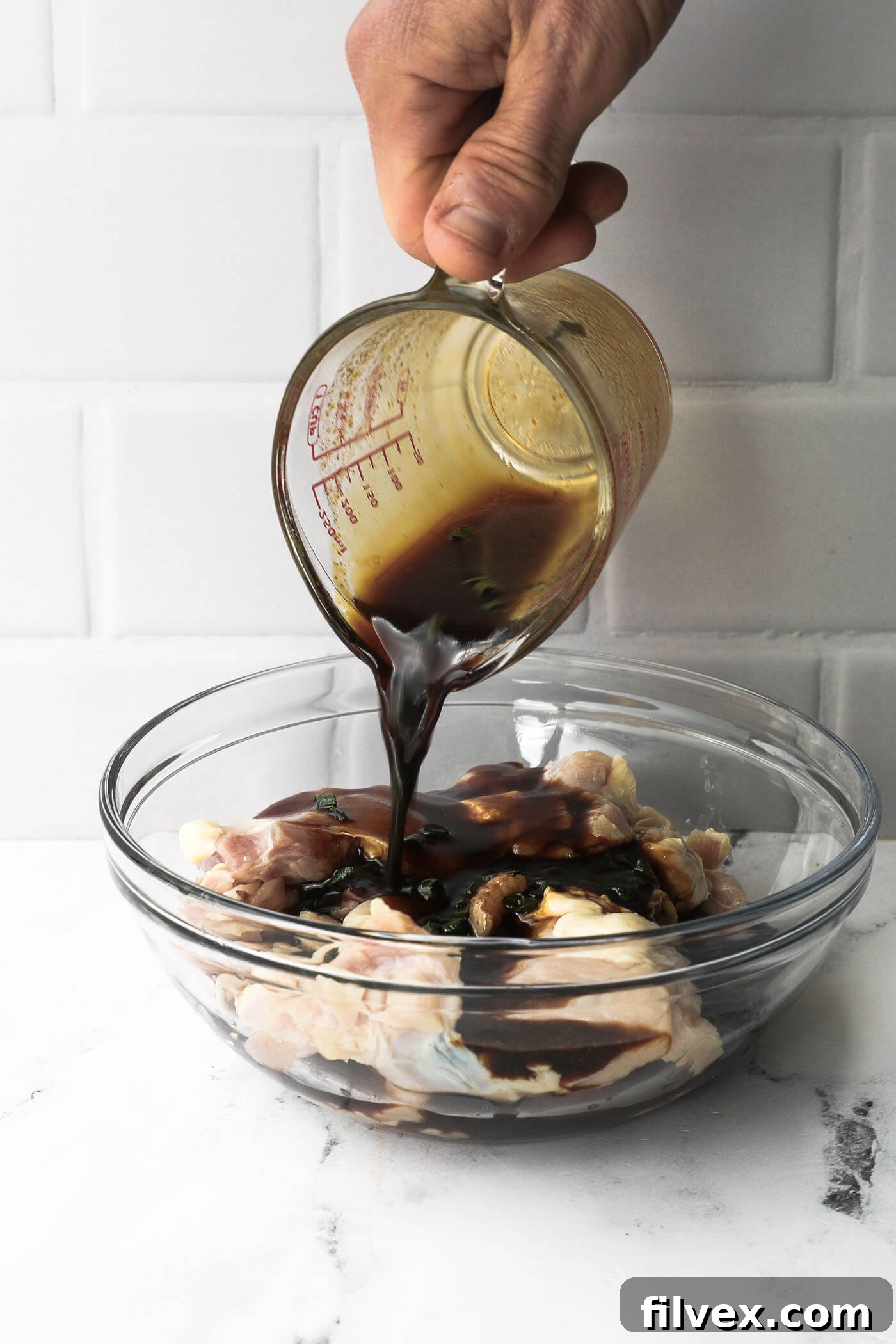 Teriyaki marinade being poured over raw chicken pieces in a clear glass bowl, illustrating the marinating process.