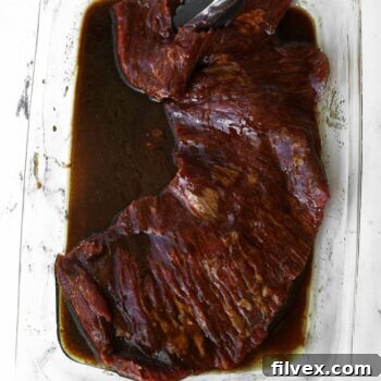 Tongs holding a piece of skirt steak dripping with balsamic marinade, ready for grilling.