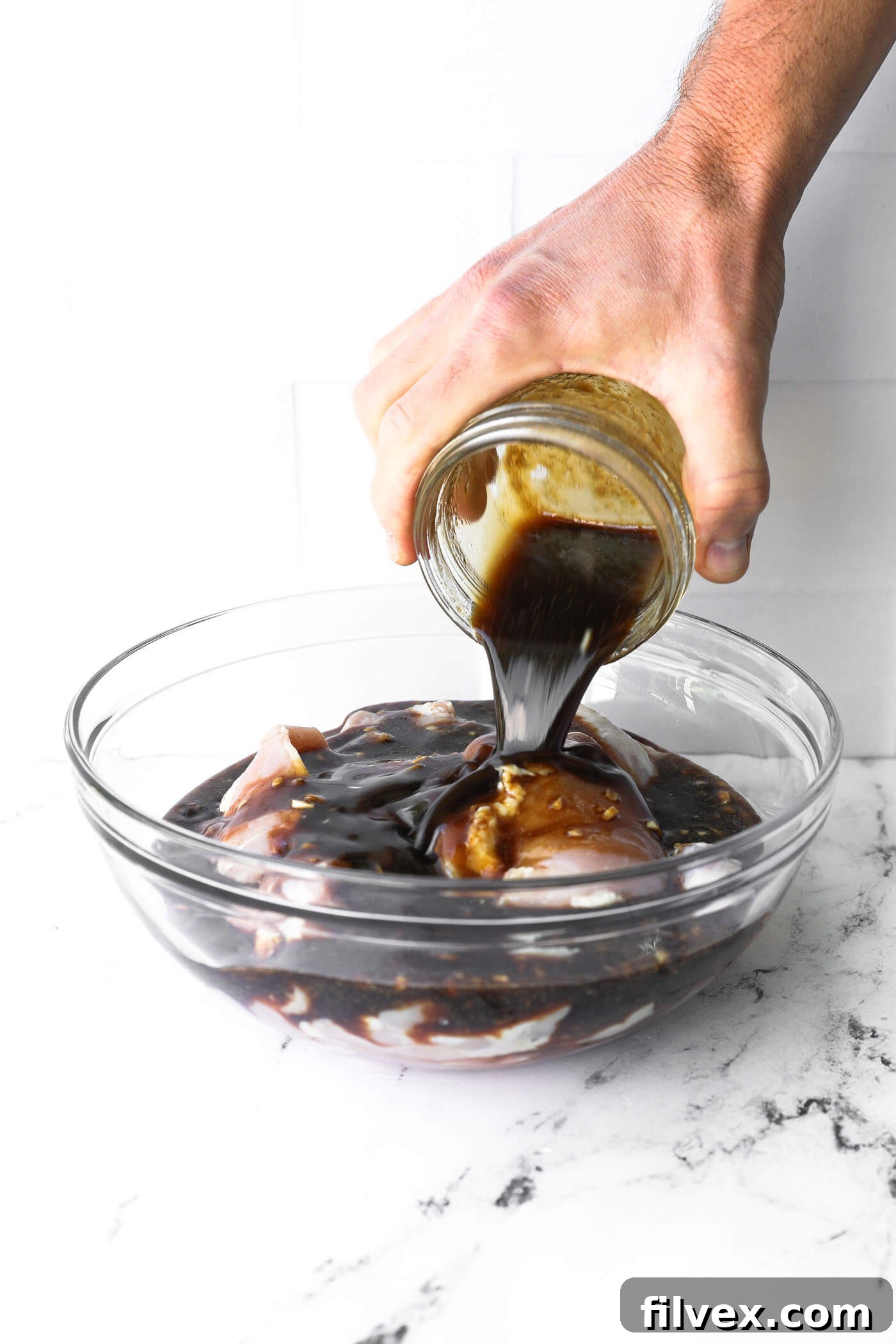Pouring honey garlic marinade over raw chicken in a glass bowl.