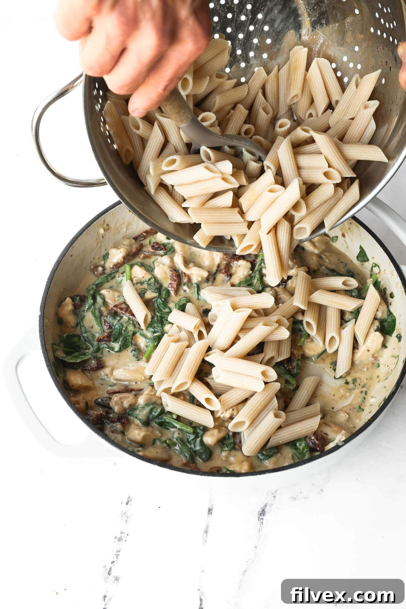 Cooked penne pasta being added to the chicken and sauce mixture in a skillet.