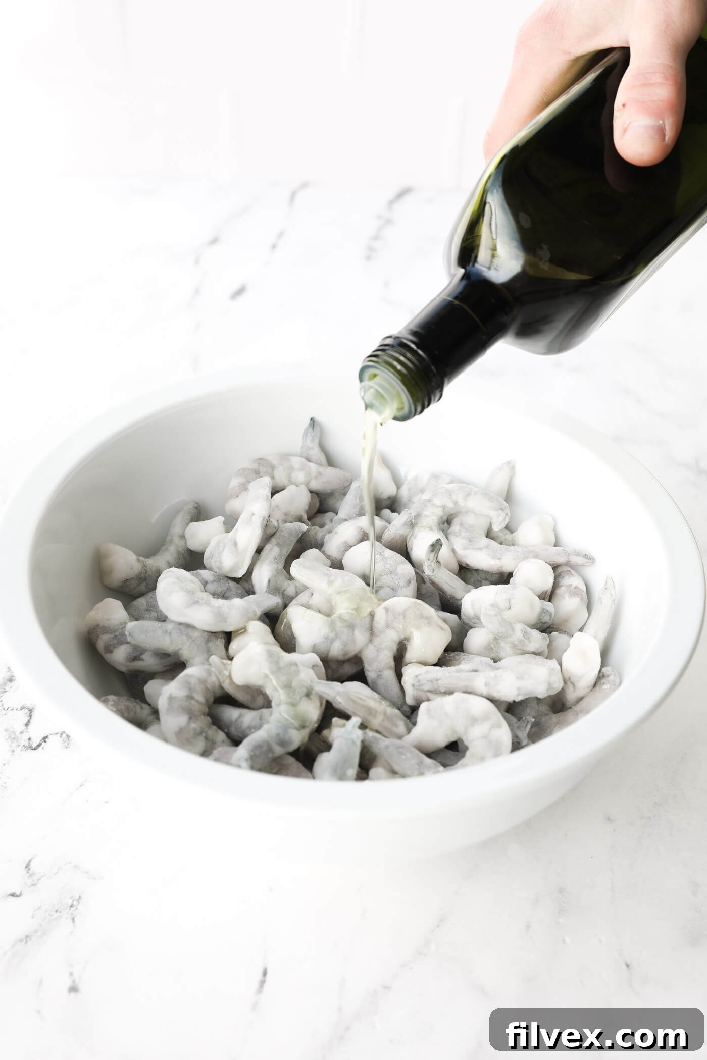 A stream of olive oil being poured over a bowl of frozen shrimp, preparing them for seasoning.