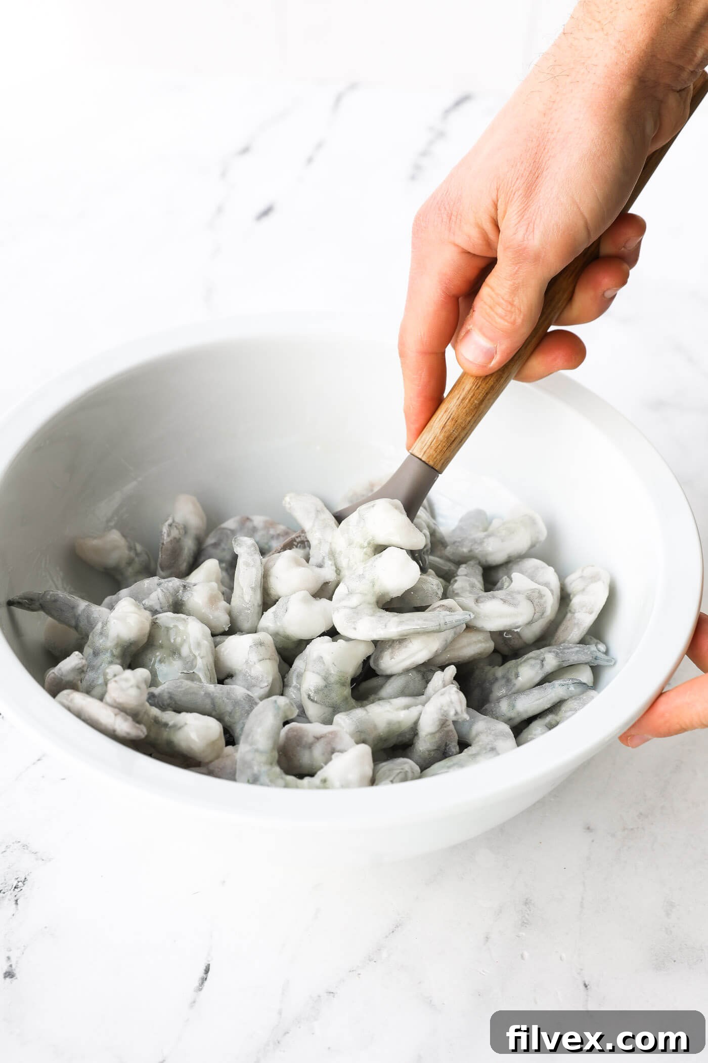 A hand gently mixing frozen shrimp with a spoon, ensuring each piece is thoroughly coated in oil.
