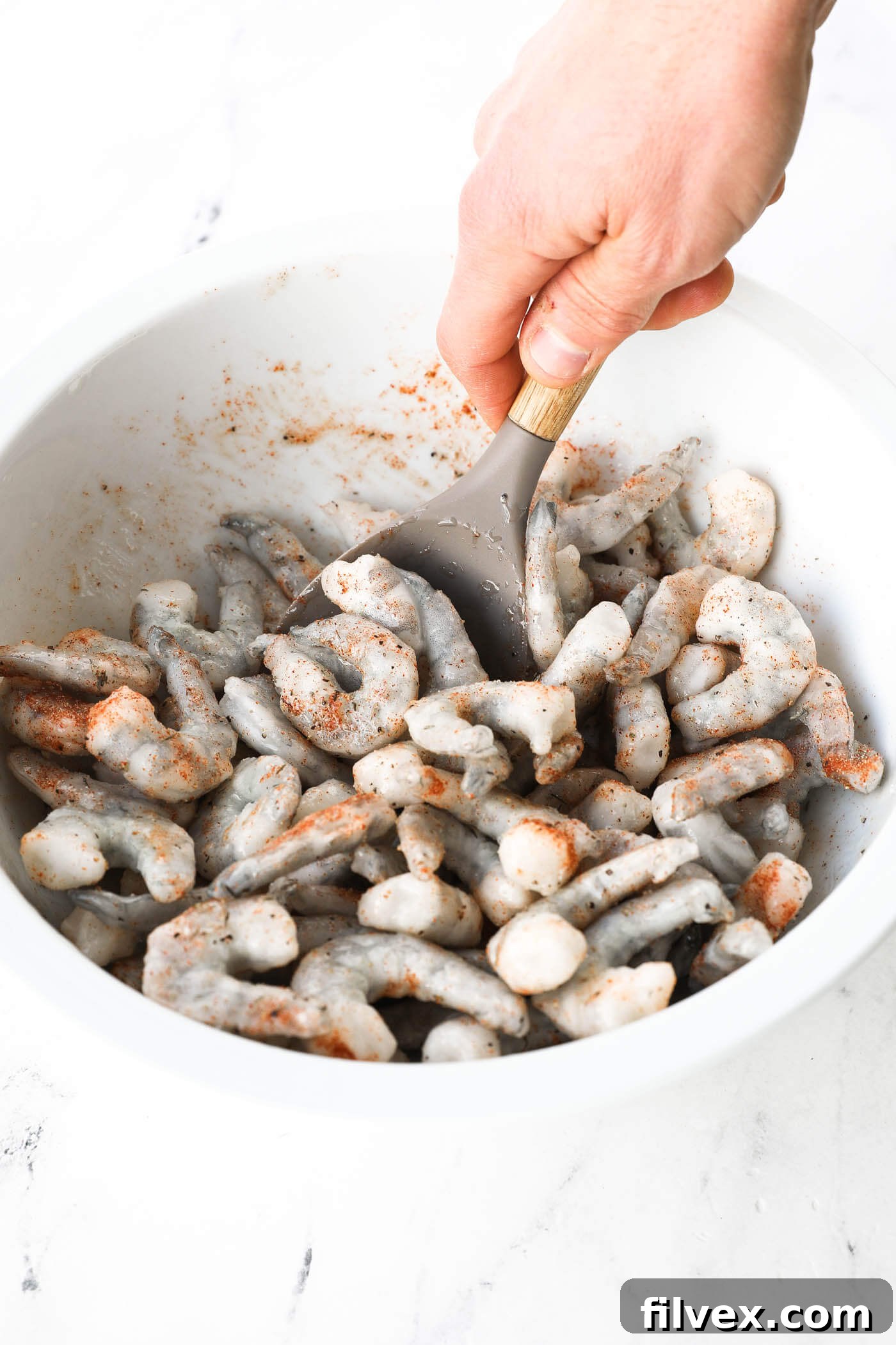 Seasoning blend being sprinkled over oiled frozen shrimp in a bowl, with a hand preparing to mix.