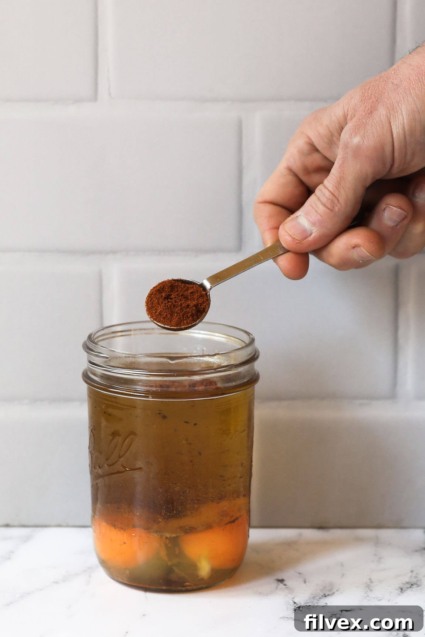 A hand pouring a teaspoon of chili seasoning into a mason jar filled with sauce ingredients.