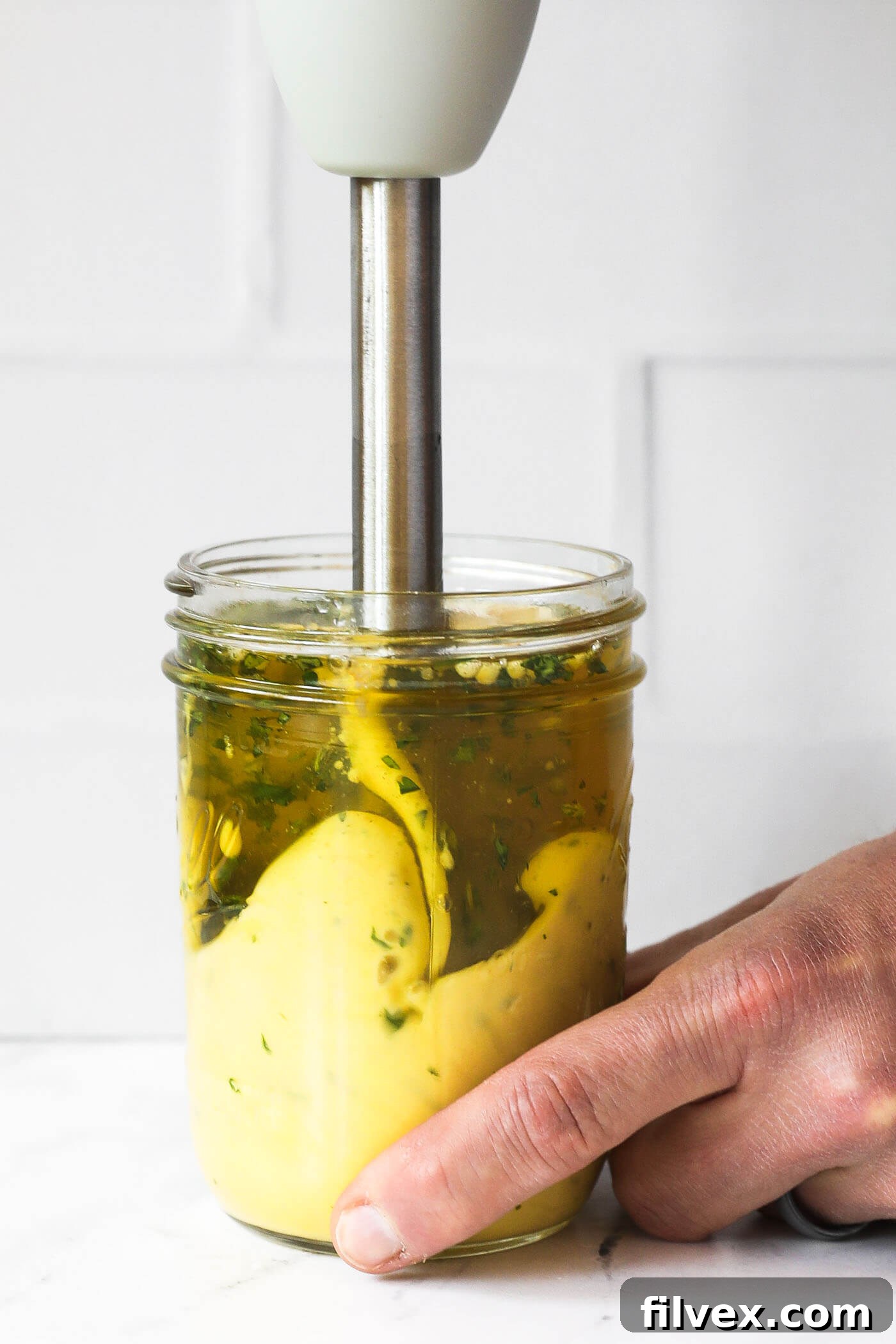 A hand holding an immersion blender at the very bottom of a mason jar, blending the cilantro lime aioli ingredients to start the emulsification process.