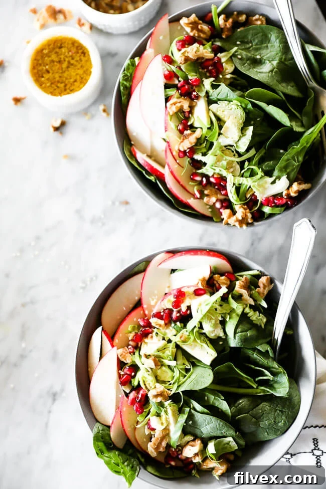 Vertical image showcasing two bowls of apple walnut spinach salad, with a small jar of dressing positioned beside them, ready for serving.