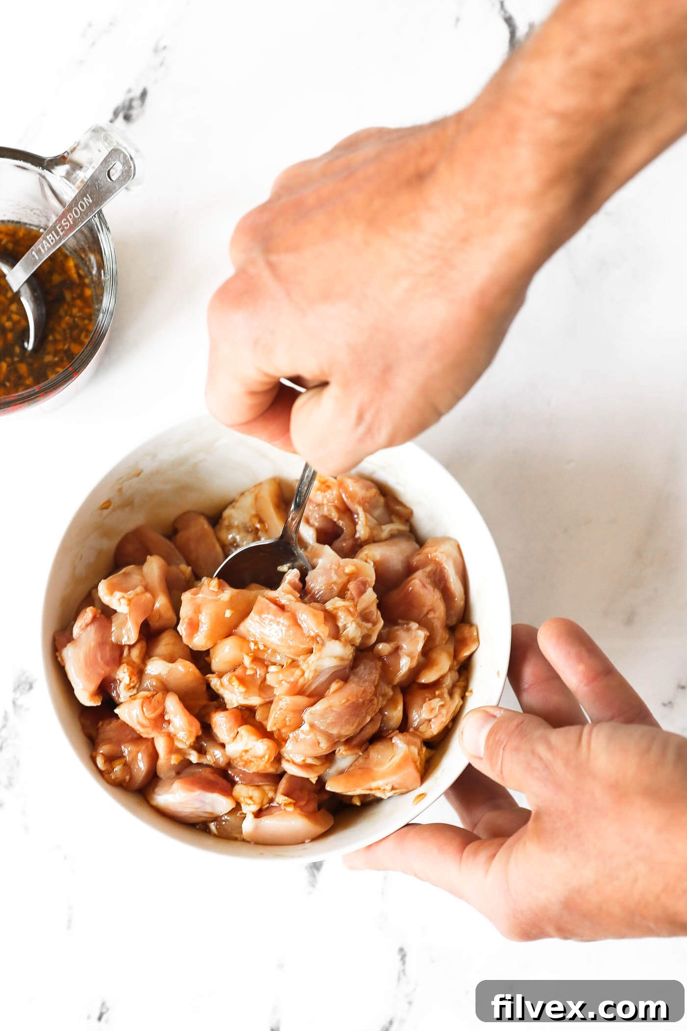 A person is holding a bowl of raw chicken pieces and mixing in a sauce.