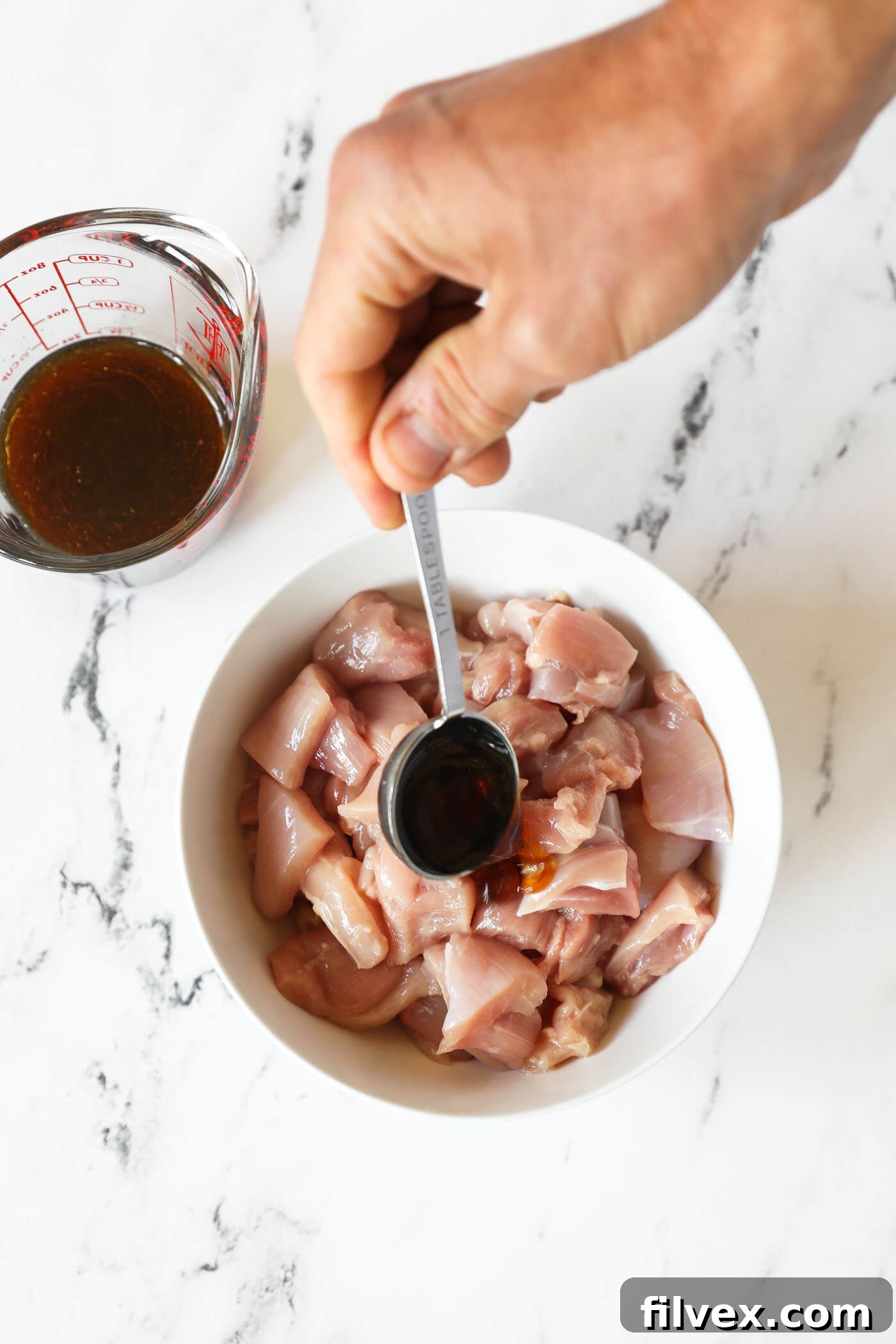 A hand spooning sauce over a bowl of raw chicken thighs cut up into bite-sized pieces for marination.