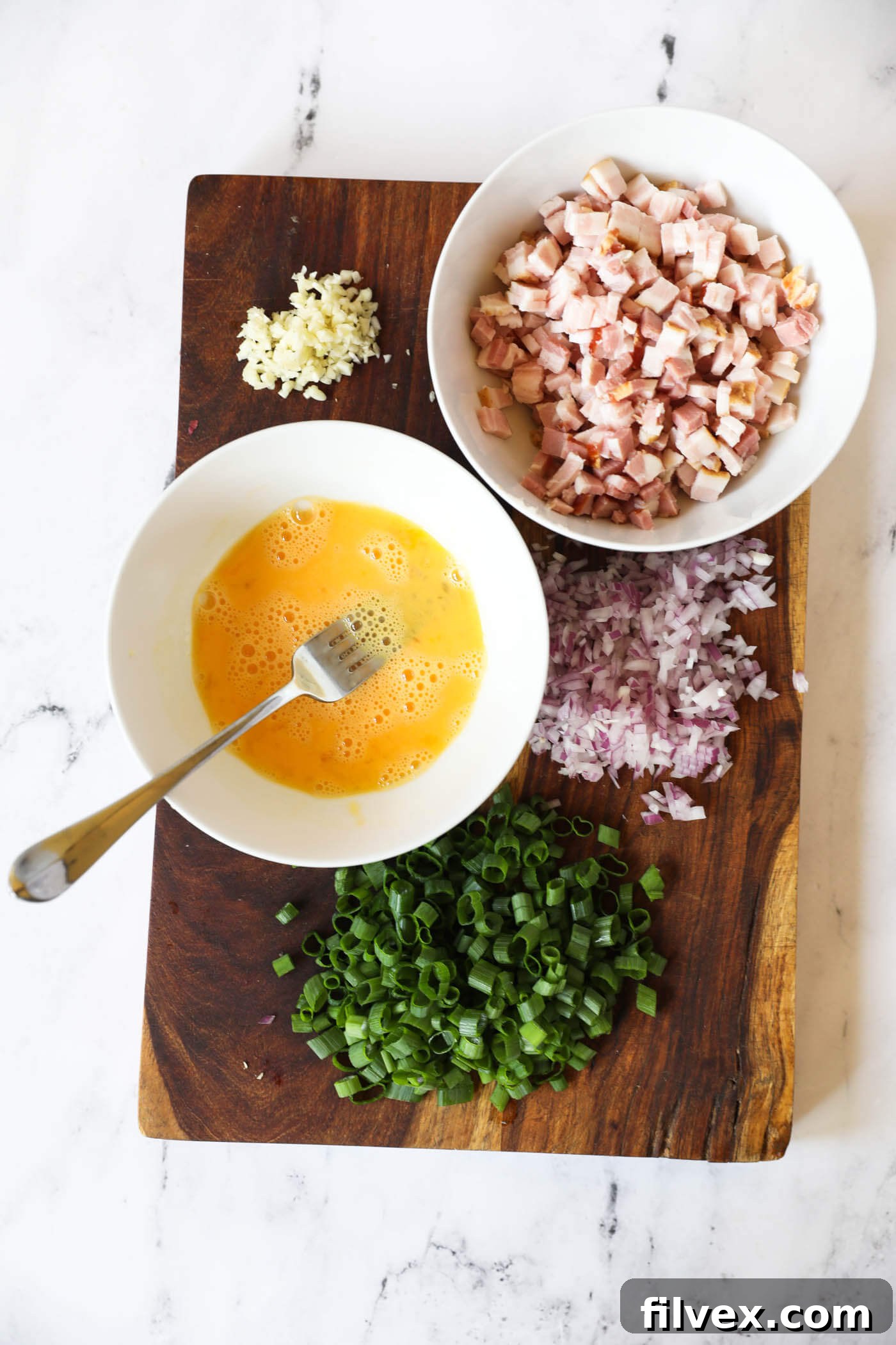 Overhead image of prepped ingredients on a cutting board - minced garlic, chopped bacon in a bowl, chopped shallot, chopped green onion and whisked egg in a bowl, ready for cooking.