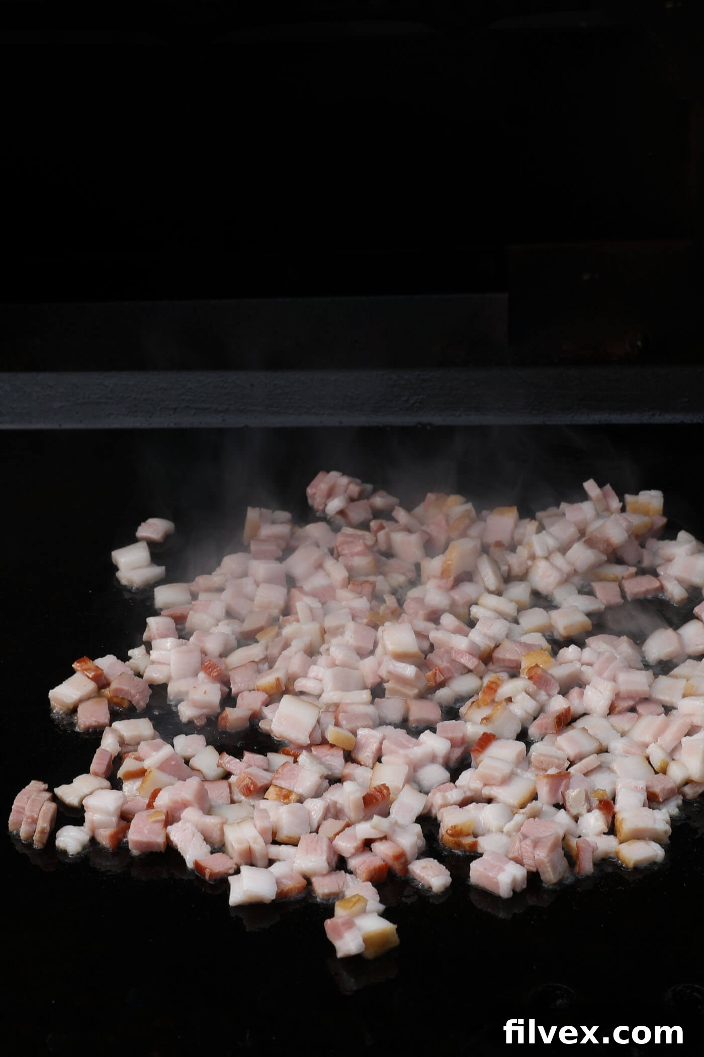 Chopped bacon cooking on a hot Blackstone griddle in a separate section, turning crispy.