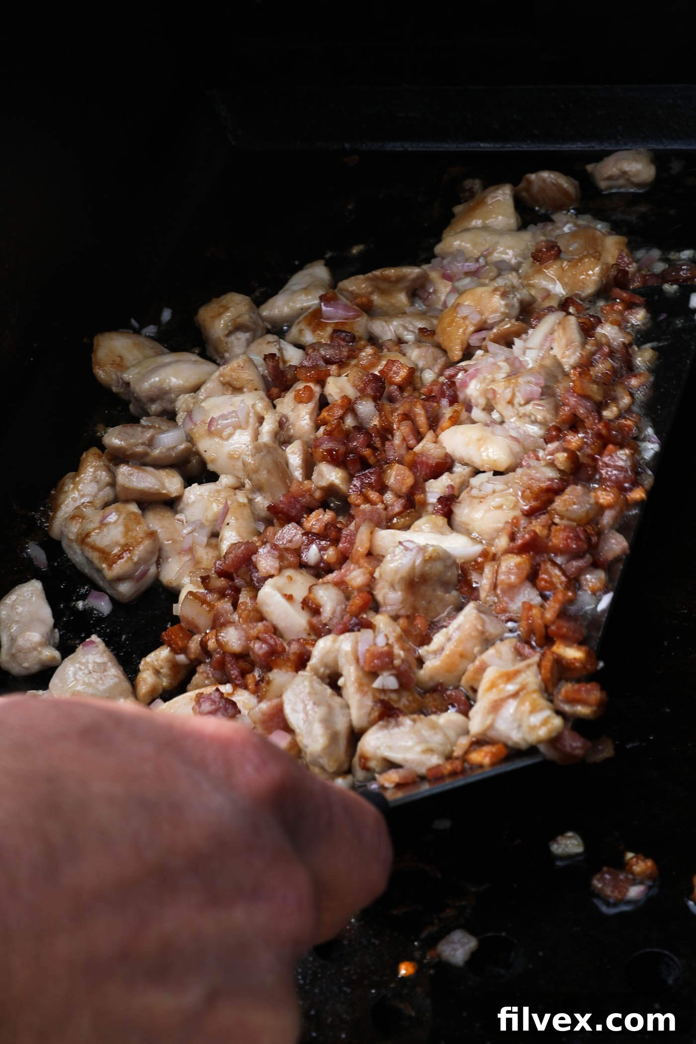 Using a large spatula to combine the chicken, garlic, shallot and bacon together on the griddle, preparing for the next step.