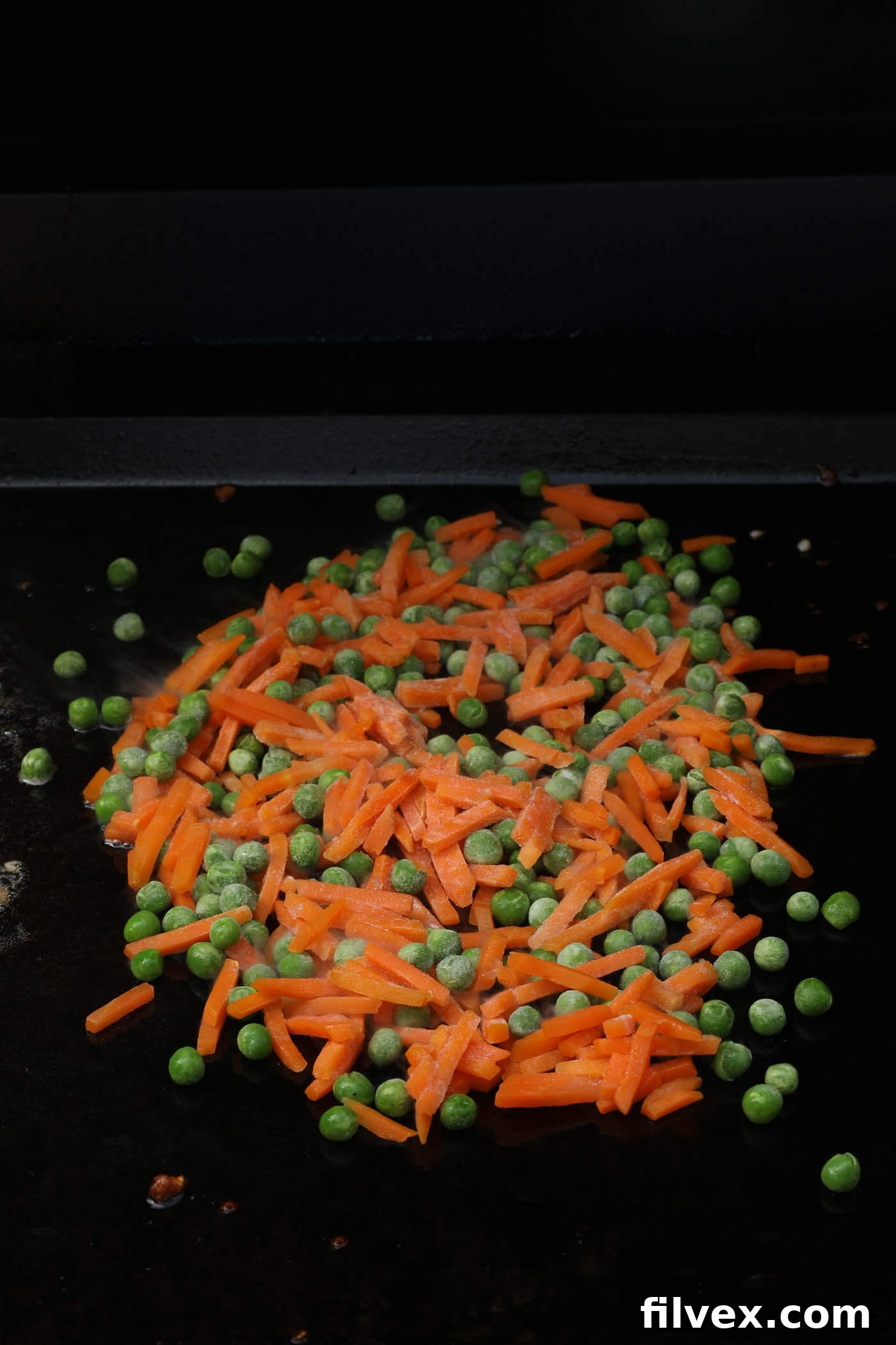 Frozen peas and carrots added to the griddle top, beginning to thaw and cook.