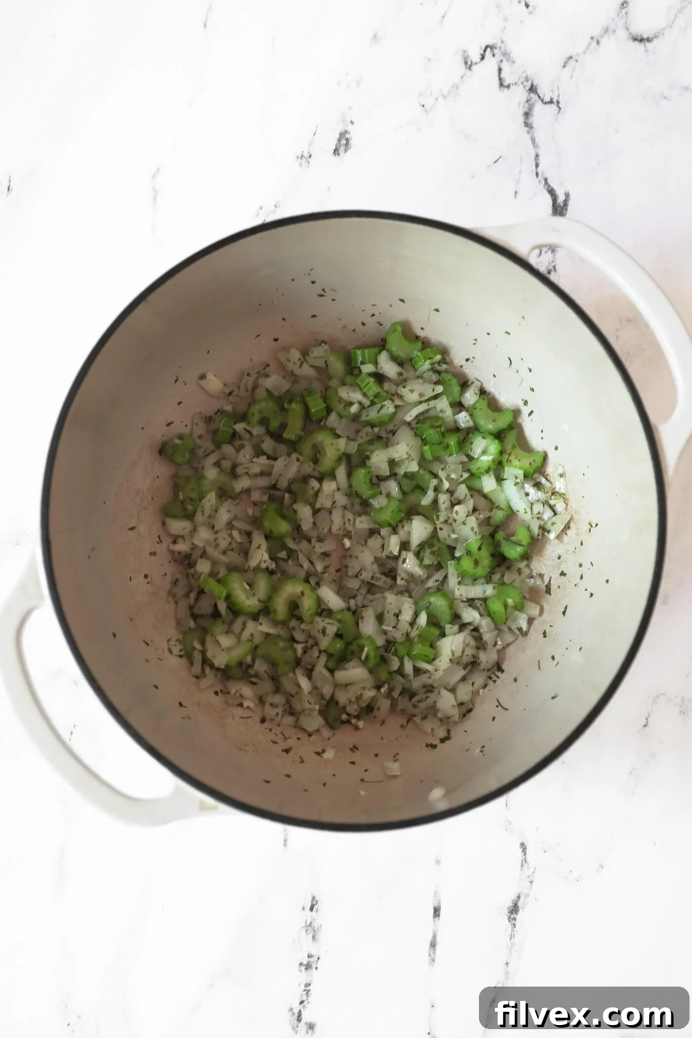 Diced onion, chopped celery, minced garlic, and aromatic Italian seasonings being sautéed in avocado oil within a Dutch oven, releasing fragrant aromas.