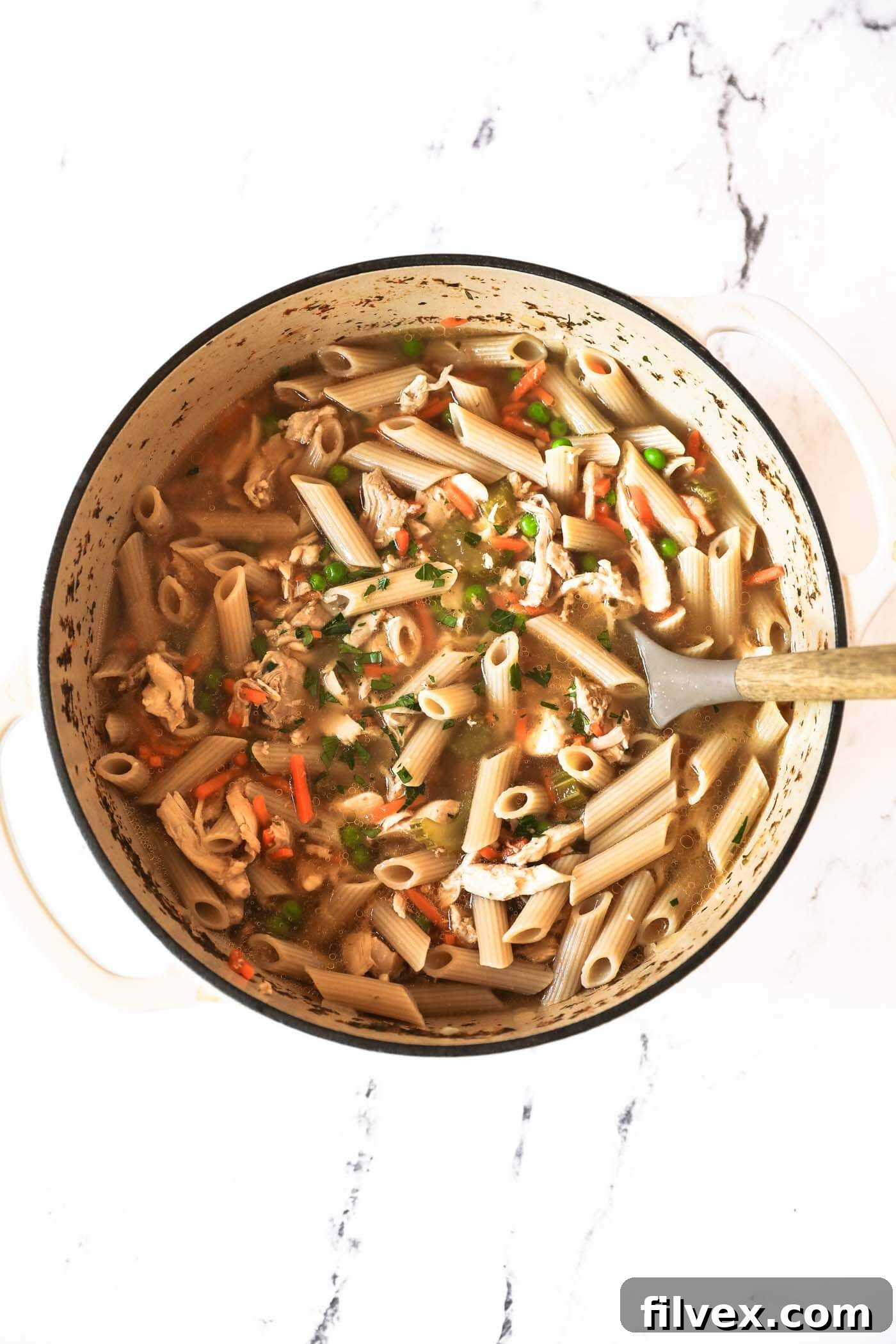 A Dutch oven filled with vibrant chicken noodle soup, with shredded chicken and frozen peas and carrots stirred back in, creating a colorful and complete meal.