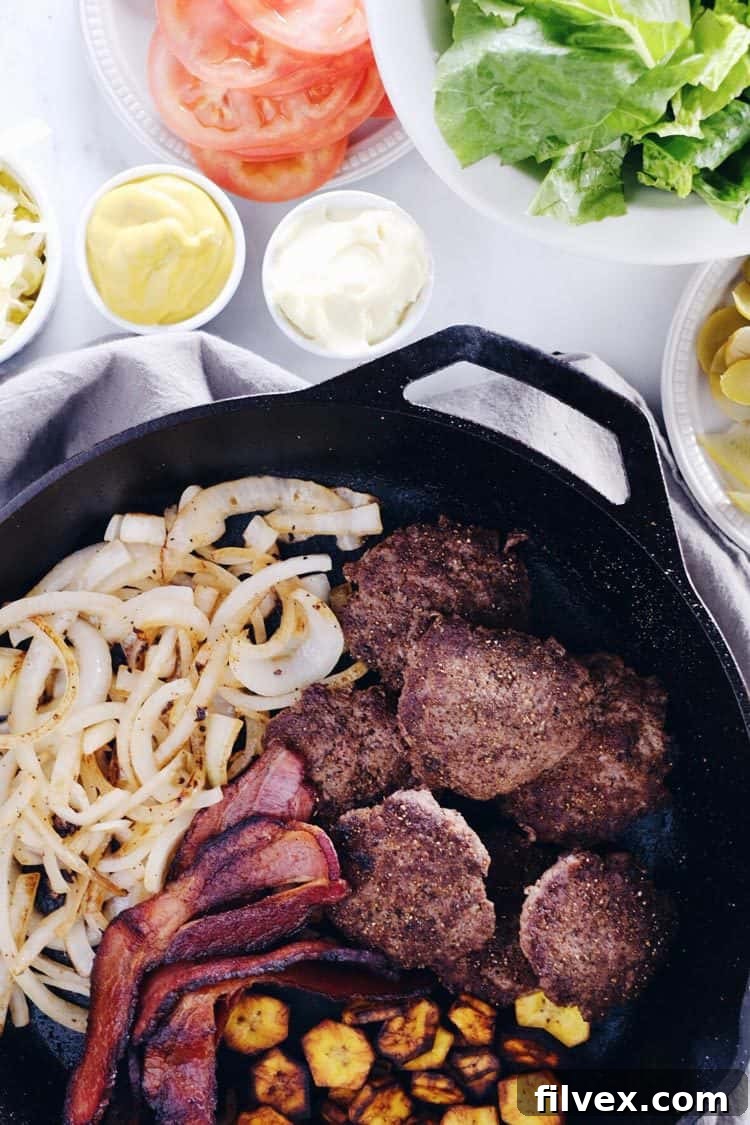 A large cast-iron skillet filled with sizzling mini burgers, crispy bacon, caramelized onions, and golden-brown pan-fried plantains, ready to be assembled into bowls.
