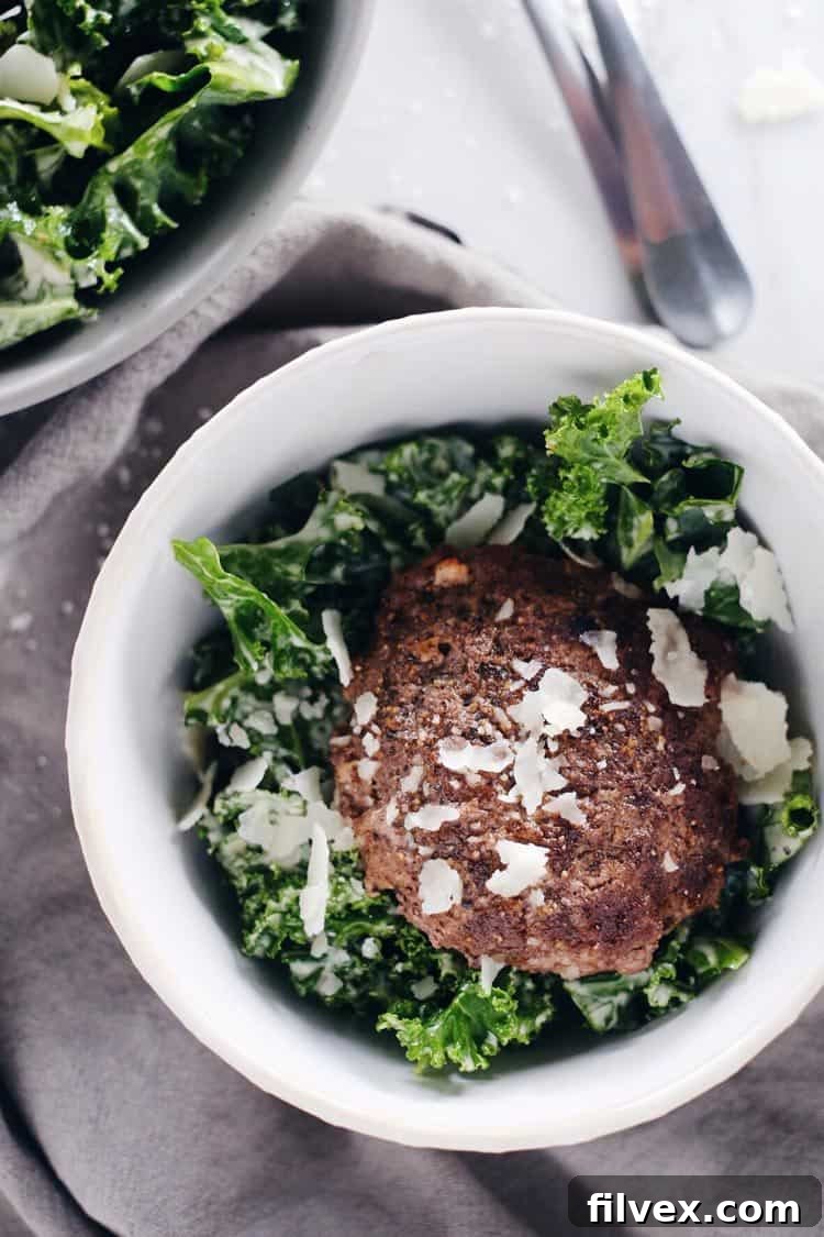 A close-up view of the hearty Kale Caesar Burger Bowl, showcasing the juicy grass-fed burger patty nestled on a bed of massaged kale with creamy Paleo Caesar dressing. Optional parmesan cheese adds a gourmet touch.
