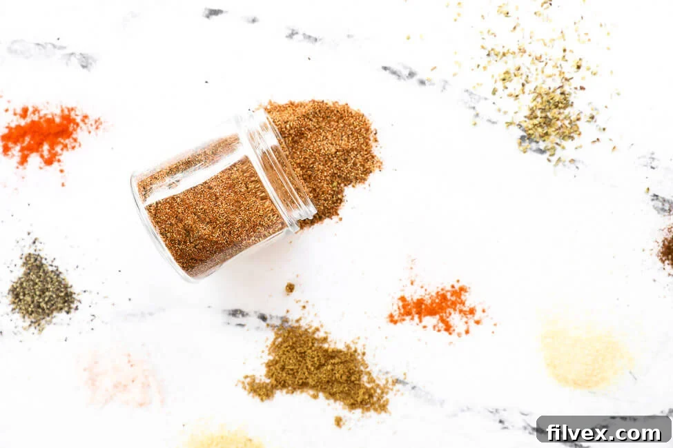 Various individual spices scattered on a light countertop, with a small, clear glass jar in the center, ready to be filled with a blend.