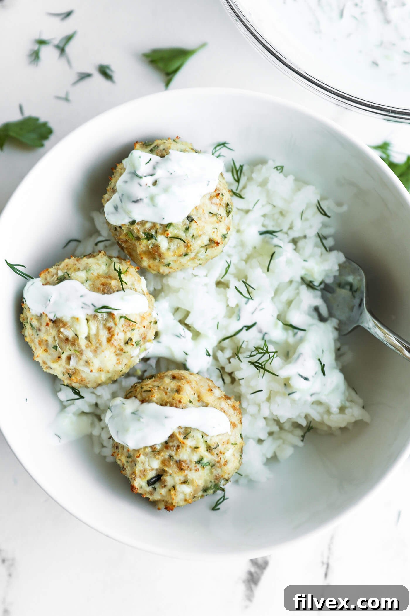 Three Greek chicken meatballs with white sauce served on a bed of rice, garnished with fresh dill, in a white bowl with a fork.