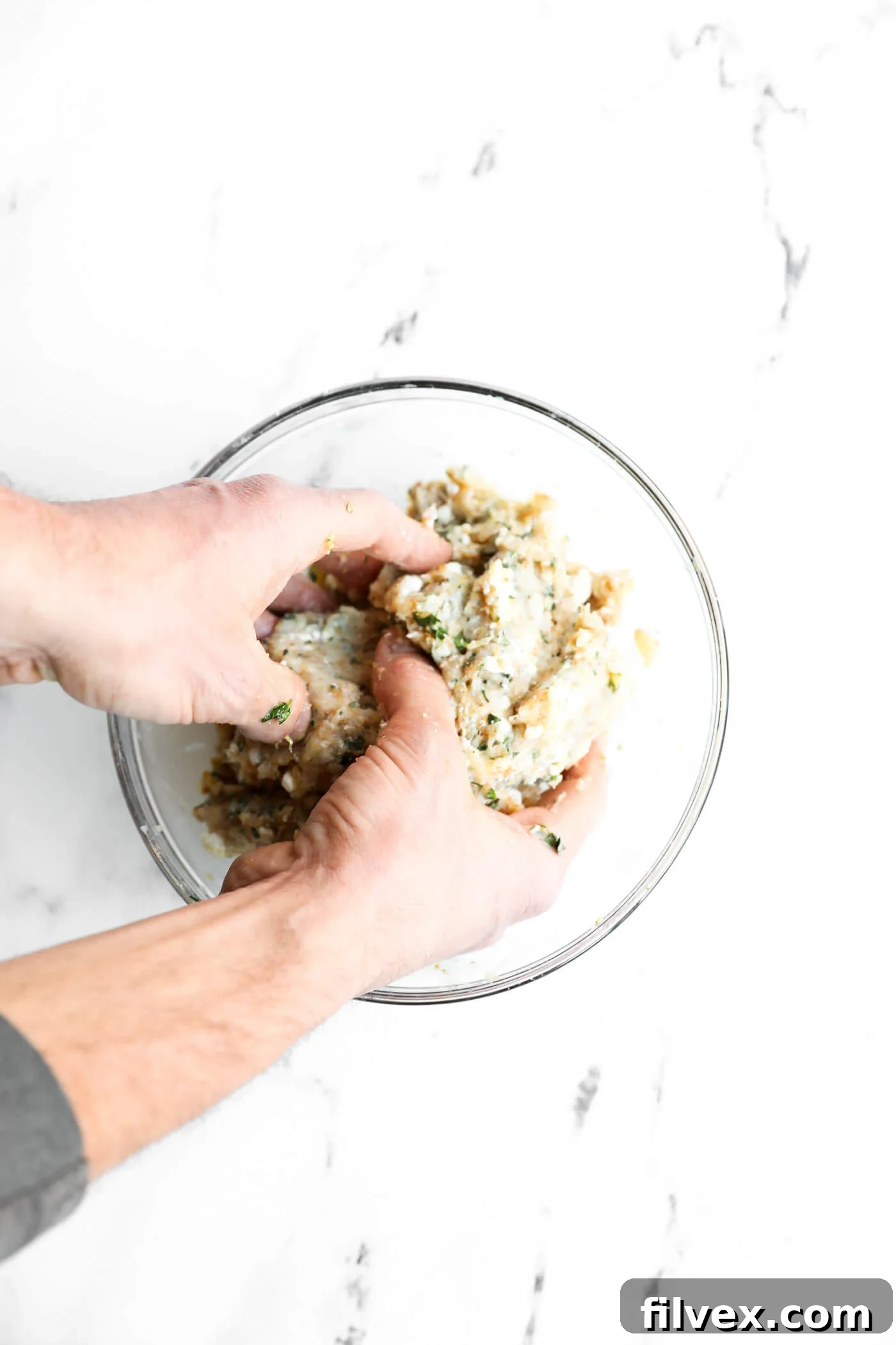 Two hands mixing a mixture of herbs, breadcrumbs, and ground meat in a clear glass bowl on a white marble surface.