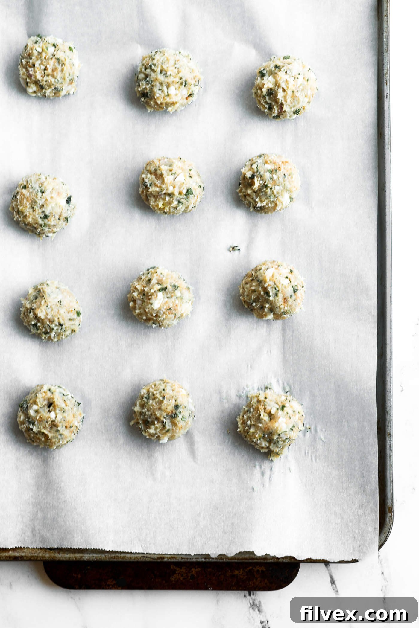 Twelve uncooked, round, breaded balls arranged on a parchment-lined baking sheet, ready to be baked.