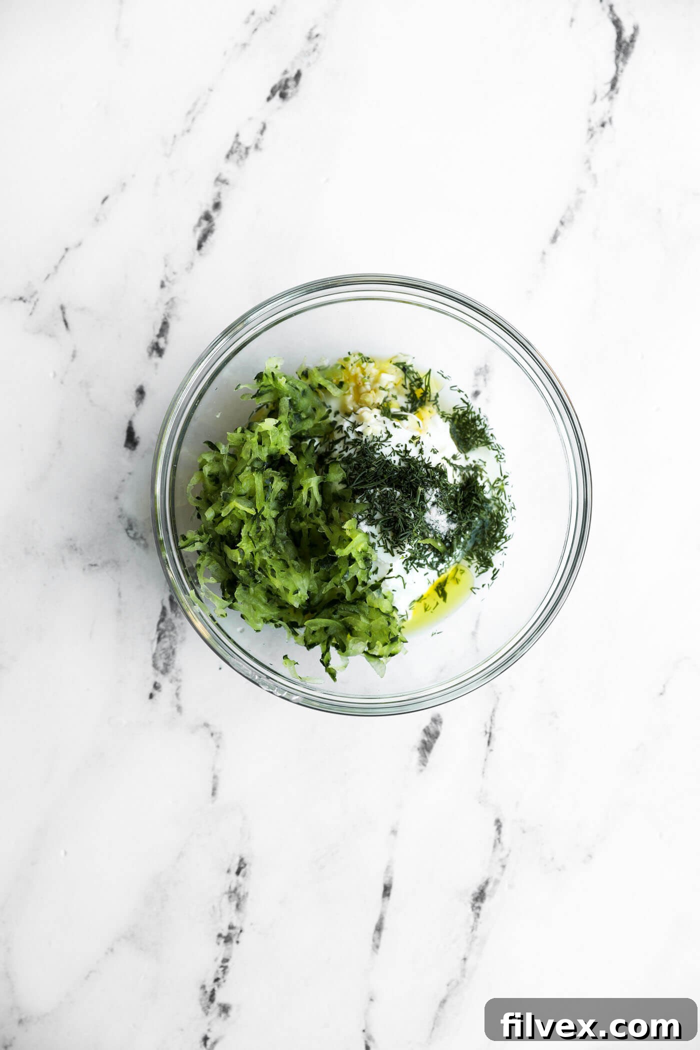 A glass bowl on a marble surface containing grated zucchini, chopped herbs, minced garlic, and yogurt or sour cream.