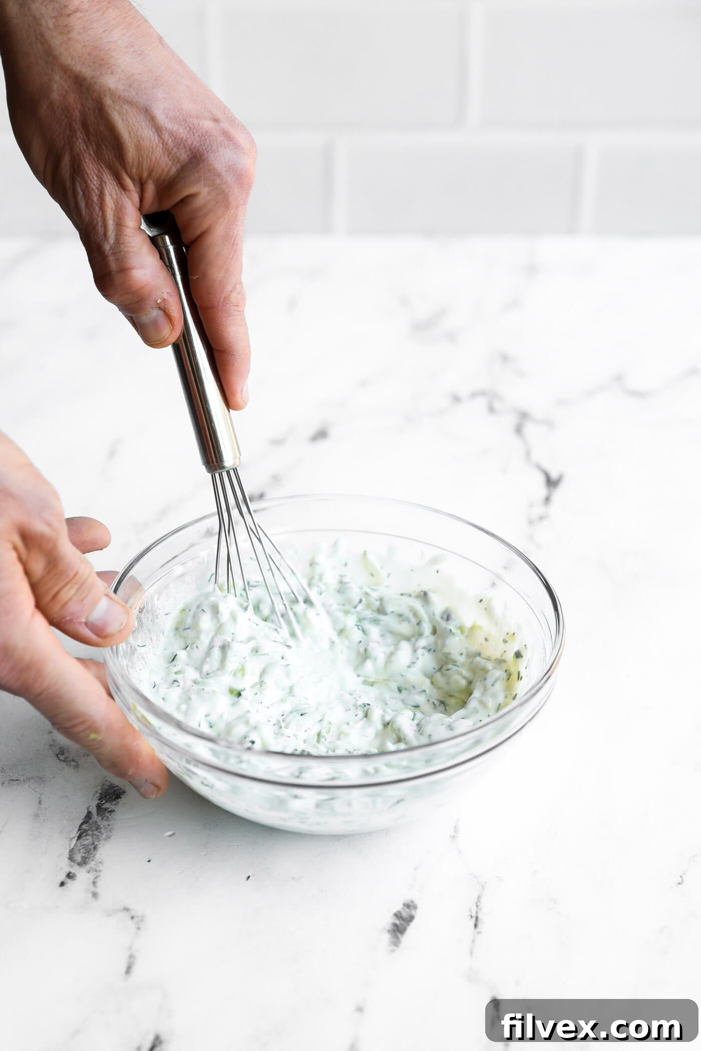 A person whisking a creamy mixture with chopped herbs in a clear glass bowl on a white marble surface.