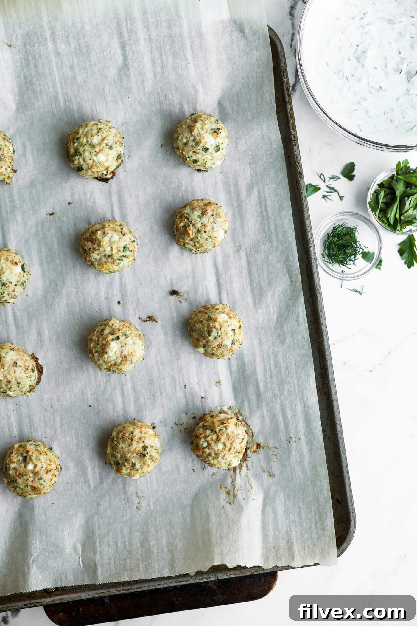 Baked herb and cheese balls on a parchment-lined baking tray, with small bowls of herbs and a bowl of white dipping sauce on the side.
