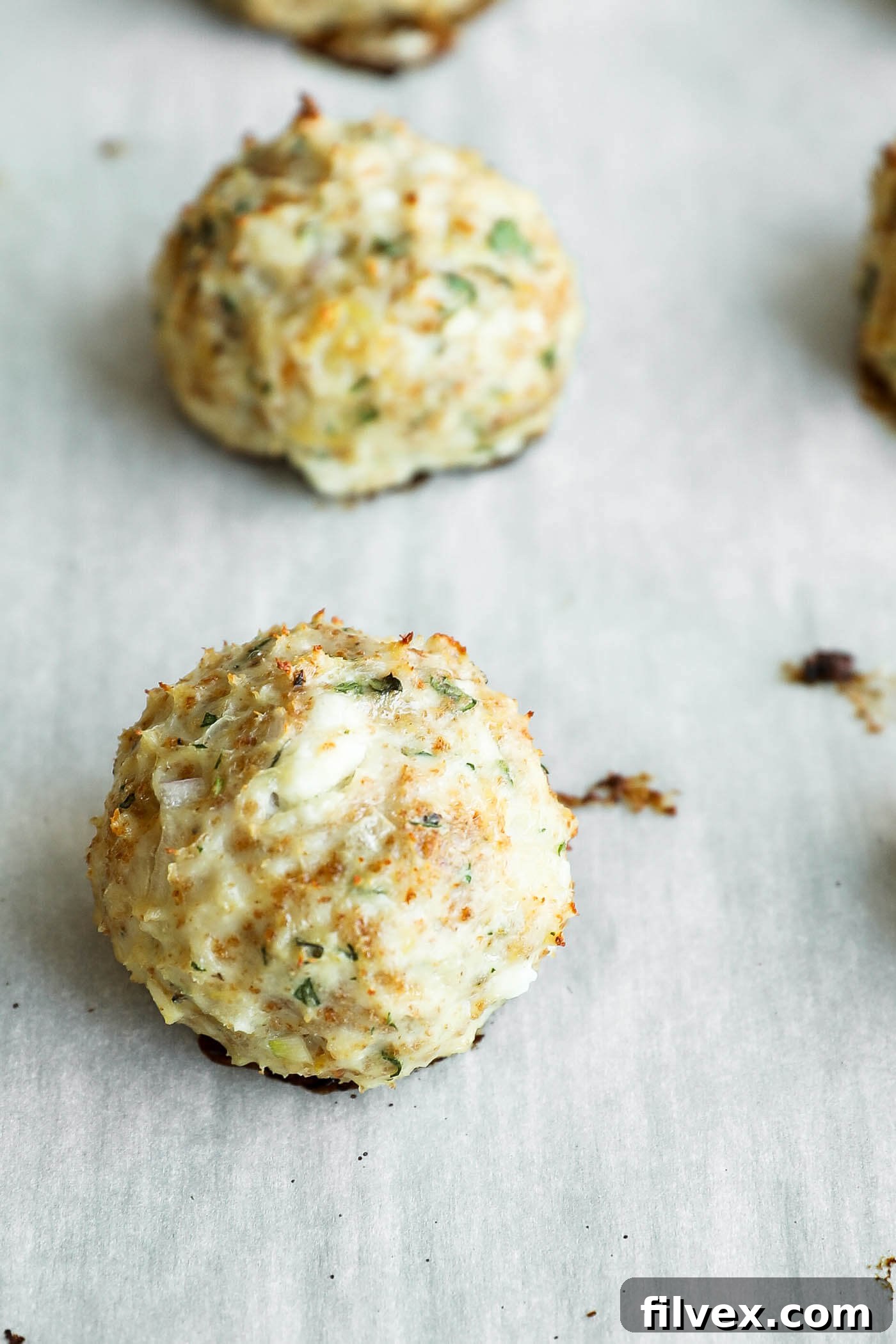 A close-up of baked, round crab cakes with herbs, resting on a parchment-lined baking sheet.