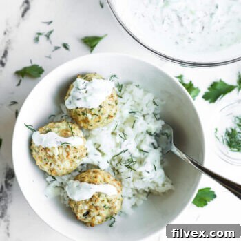 A white bowl with three meatballs on a bed of rice, topped with white sauce and herbs, with a spoon and a bowl of extra sauce beside it on a marble surface.